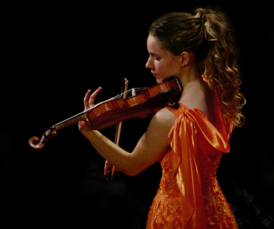 A violinist wearing an orange gown performing with the listening audience darkened out in the foreground.