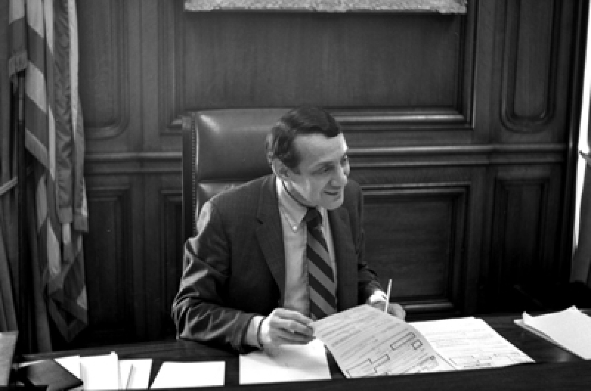 Person wearing suit seated behind a desk in a talk-backed chair with a wood-paneled wall, framed artwork, and flag alongside.