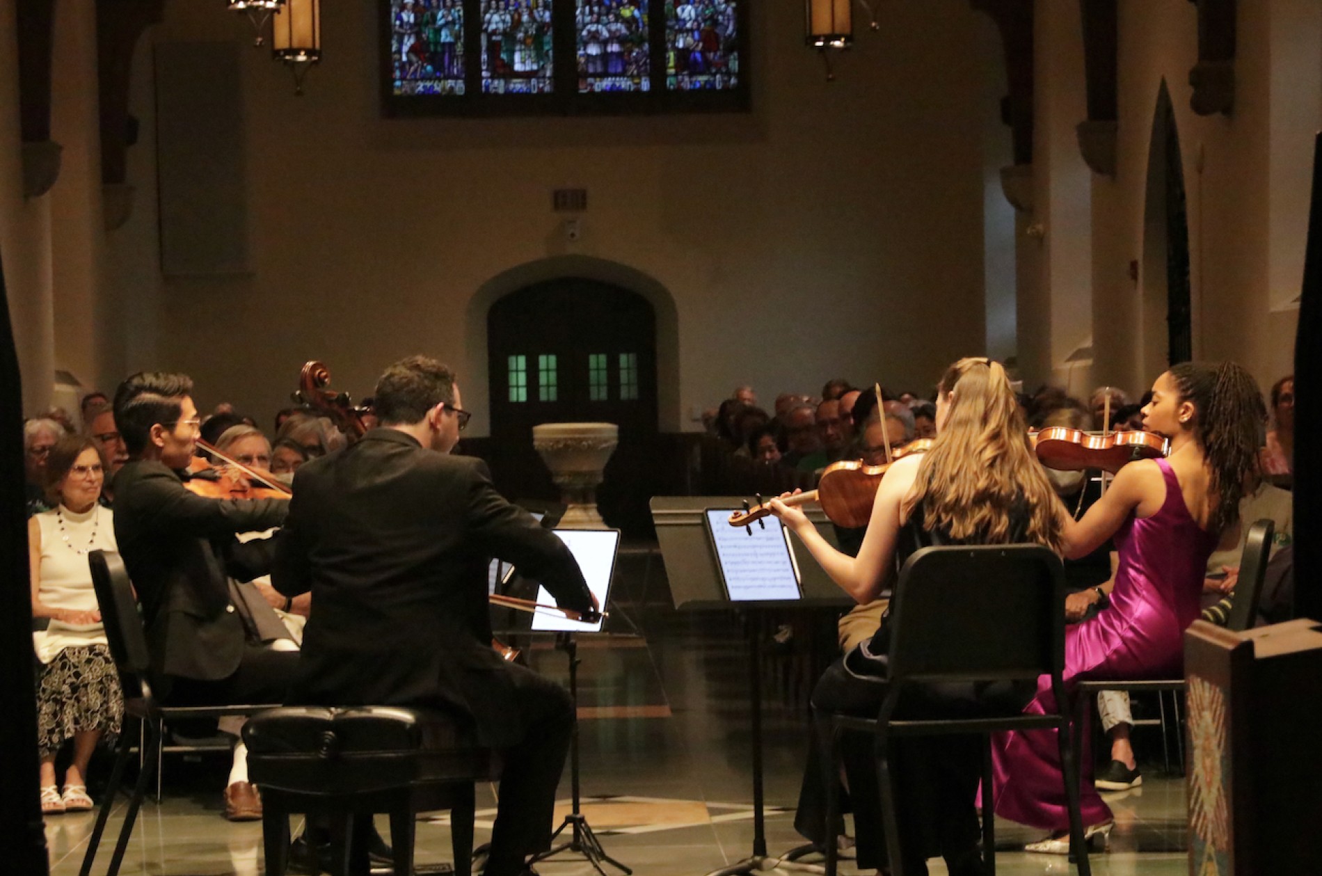 A string quartet performing in a church setting, seated at music stands with an audience seated in pews beyond.