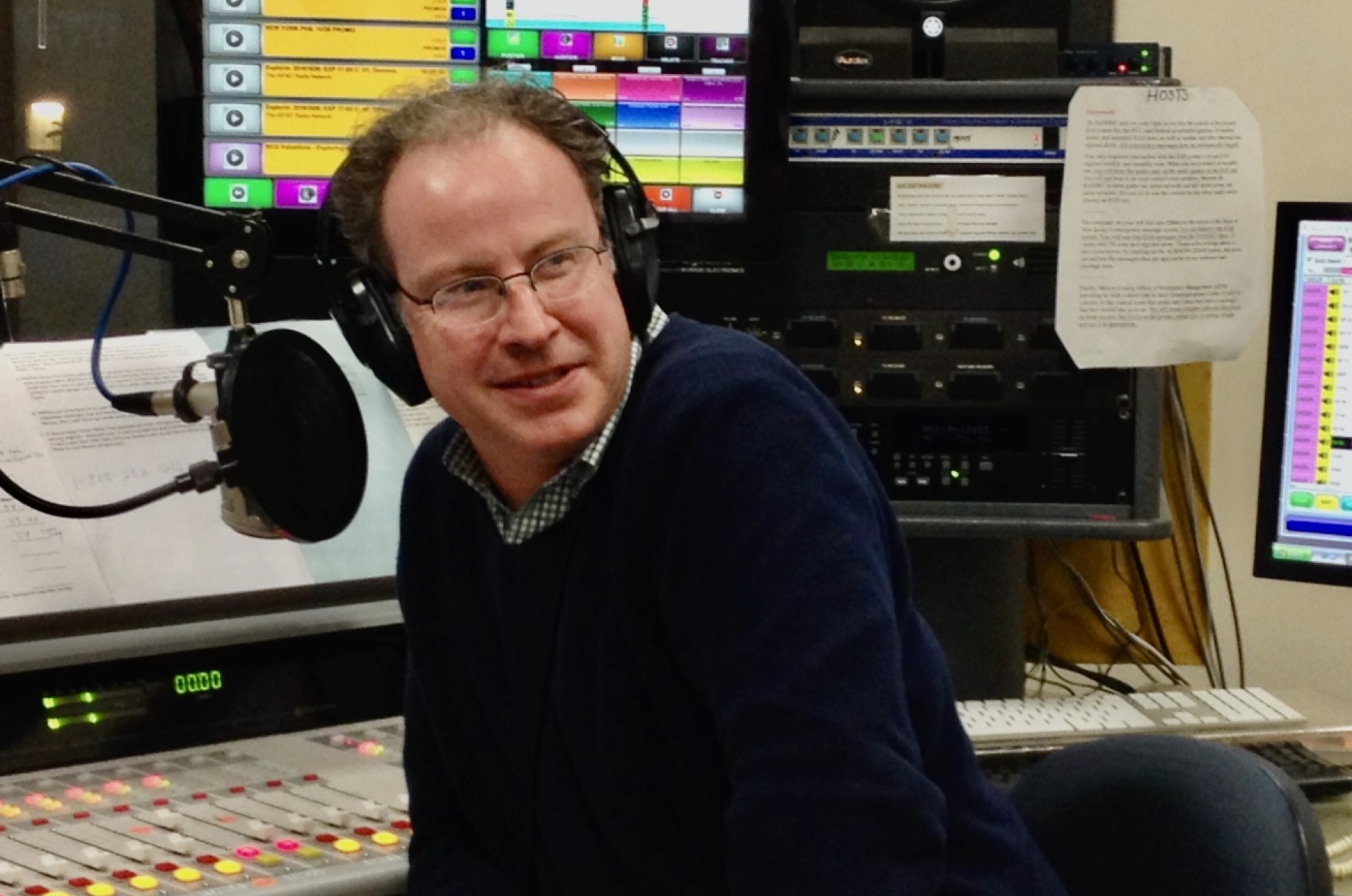 Radio host in a studio wearing headphones at a soundboard, next to a hanging microphone.