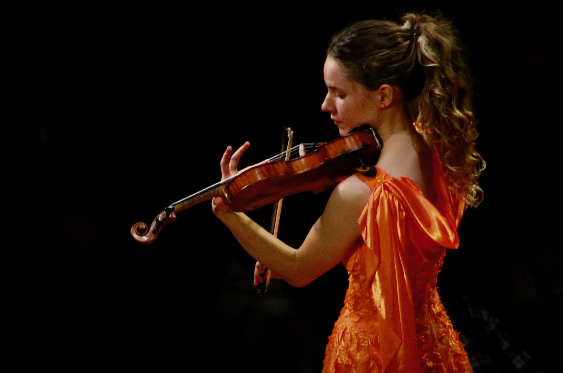 Aubrey Oliverson, violin - performing in a darkened concert hall in an orange gown - Sept 2024