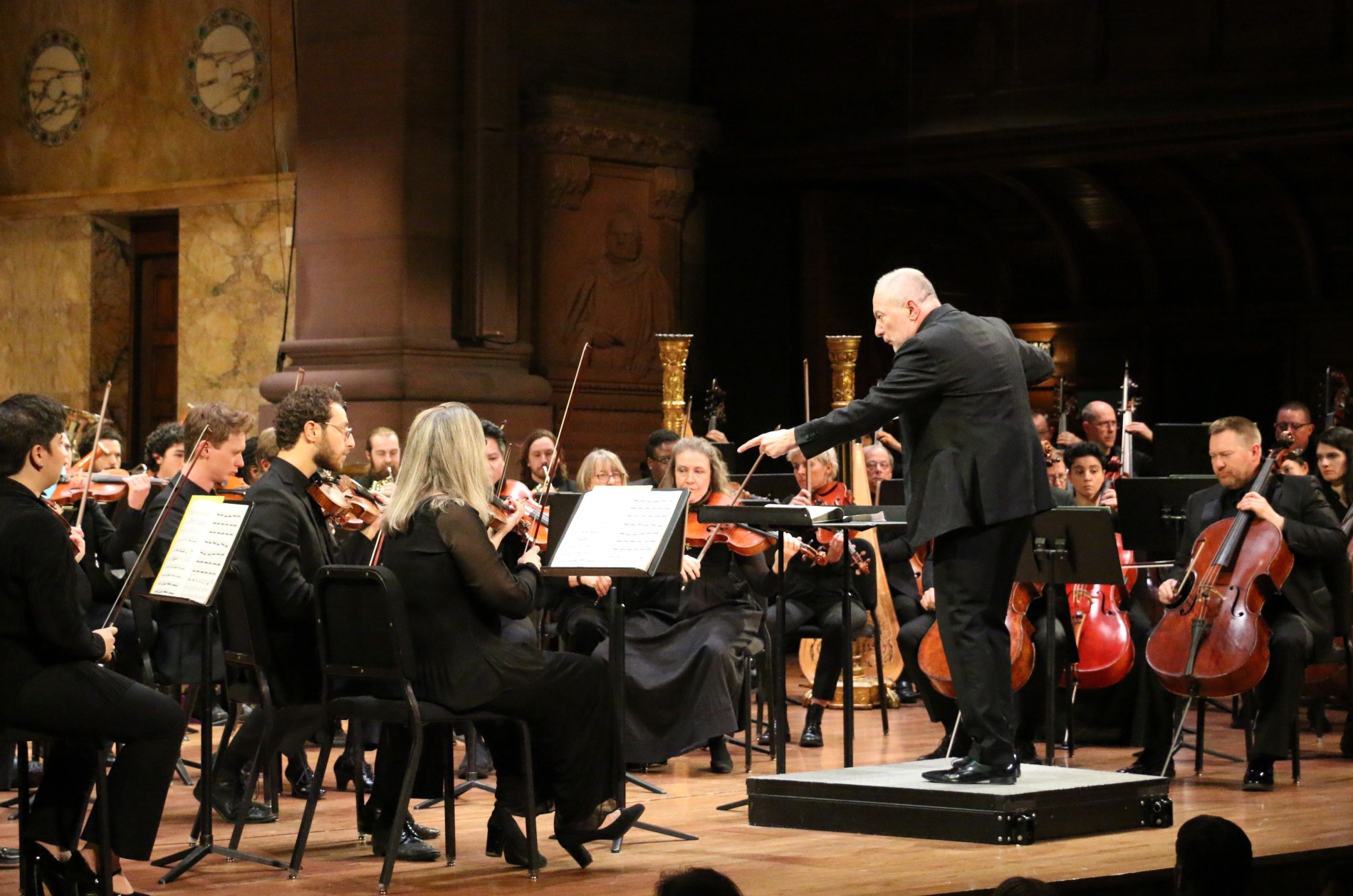 Conductor pointing wand towards violins during a performance with full orchestra.