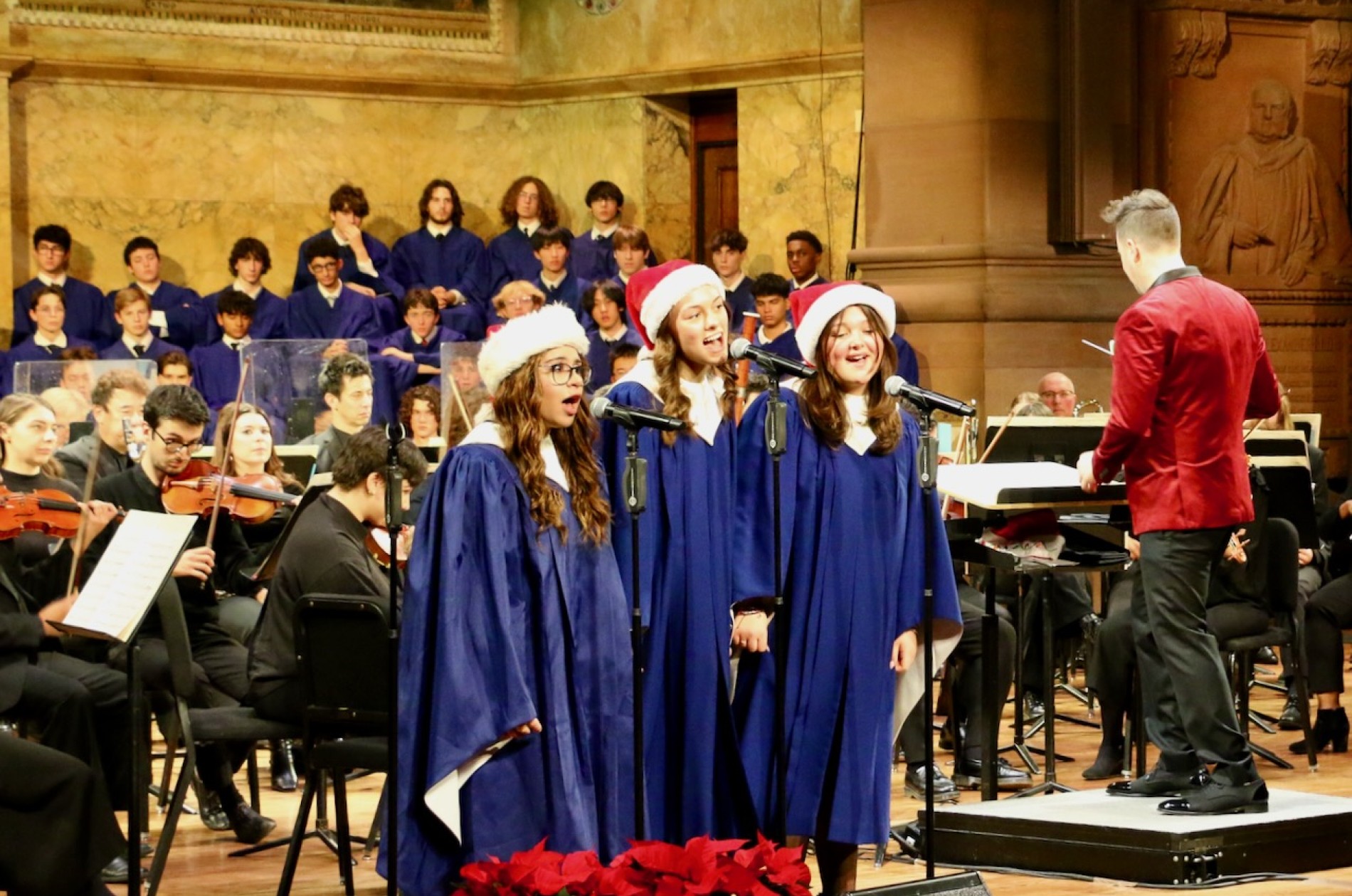 Three choir members in blue choir robes and red Santa hats singing on stage as a trio with conductor, a few orchestra members, and the full choir in the background.